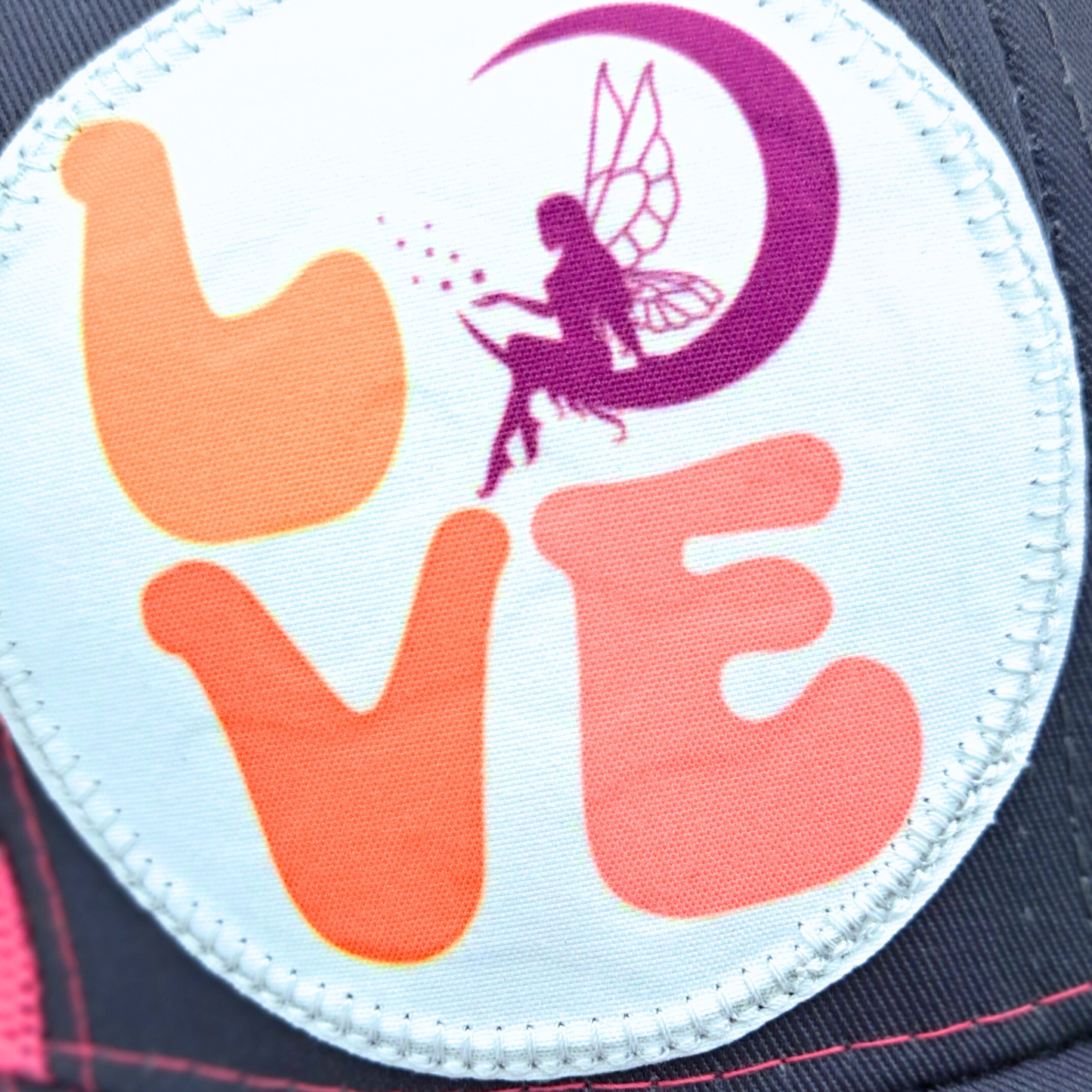 Up close view of the Large round white patch is on the right side of the hat. Patch has the word Love in bubble letters. The o is a crescent moon with a fairy sitting inside it.