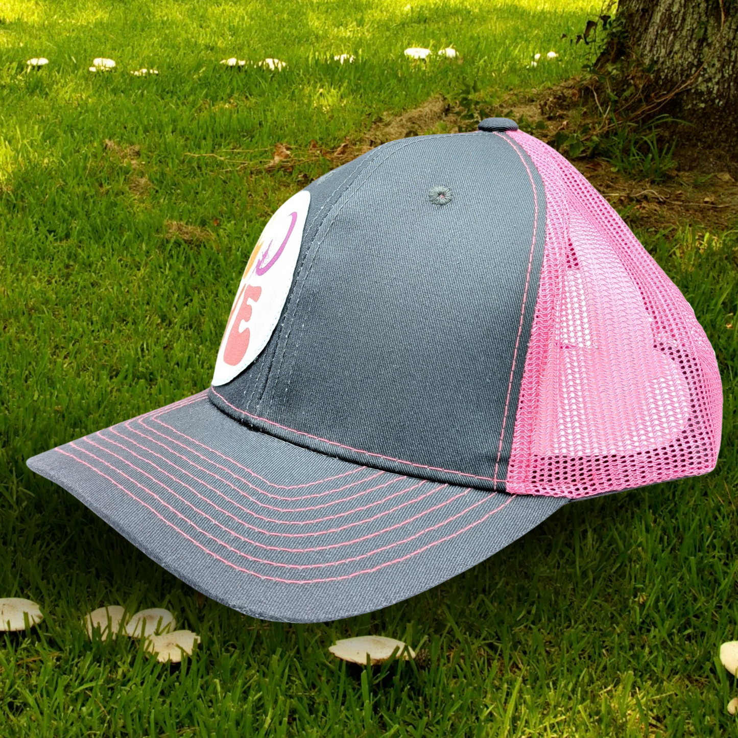 Left front view of Grey and Pink Trucker hat with hot pink stitching on the bill. Front and bill of hat are grey and the back is hot pink mesh. Large round white patch is on the right side of the hat. Patch has the word Love in bubble letters. The o is a crescent moon with a fairy sitting inside it.