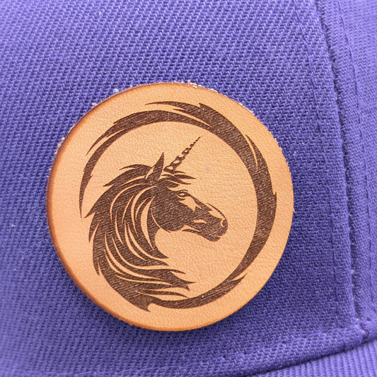 Up close view of the round tan leather patch. Laser engraved on the patch is a the detailed head of a unicorn facing into the patch. From the unicorn's mane a line swirls out framing the unicorn in a circle that ends at the top of the unicorn's head at the mane.