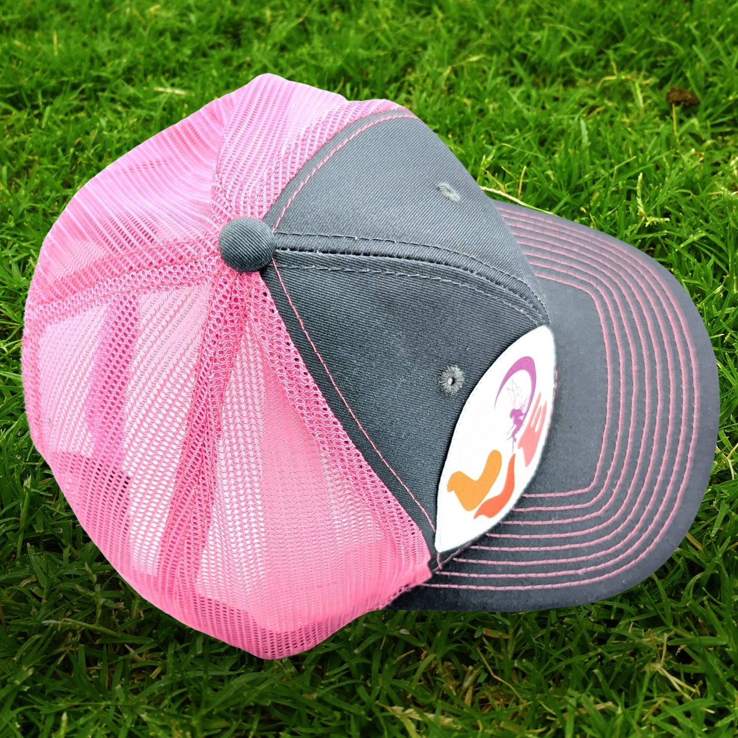 Top view of the Grey and Pink Trucker hat with hot pink stitching on the bill. Front and bill of hat are grey and the back is hot pink mesh. Large round white patch is on the right side of the hat. Patch has the word Love in bubble letters. The o is a crescent moon with a fairy sitting inside it.