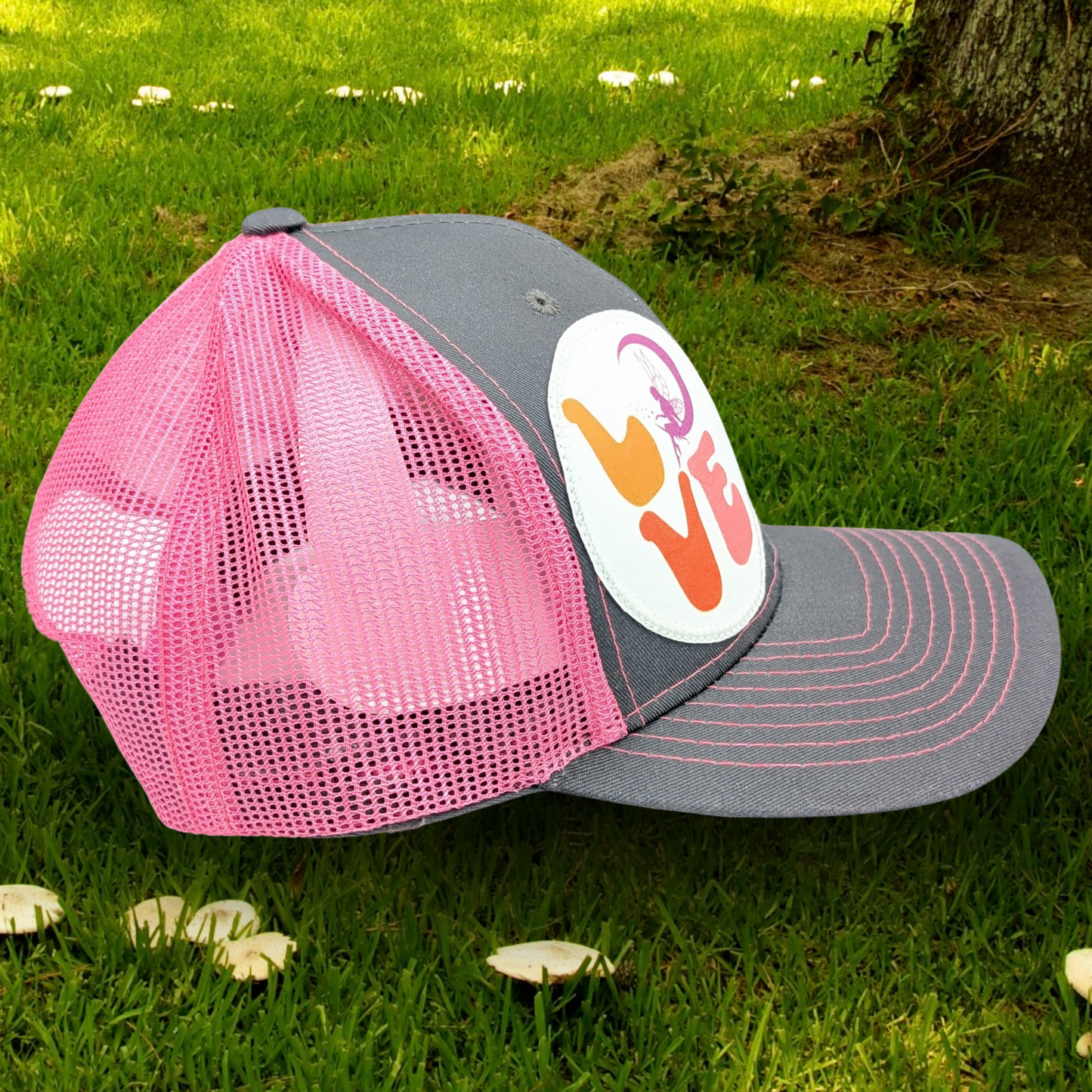 Right side view of Grey and Pink Trucker hat with hot pink stitching on the bill. Front and bill of hat are grey and the back is hot pink mesh. Large round white patch is on the right side of the hat. Patch has the word Love in bubble letters. The o is a crescent moon with a fairy sitting inside it.