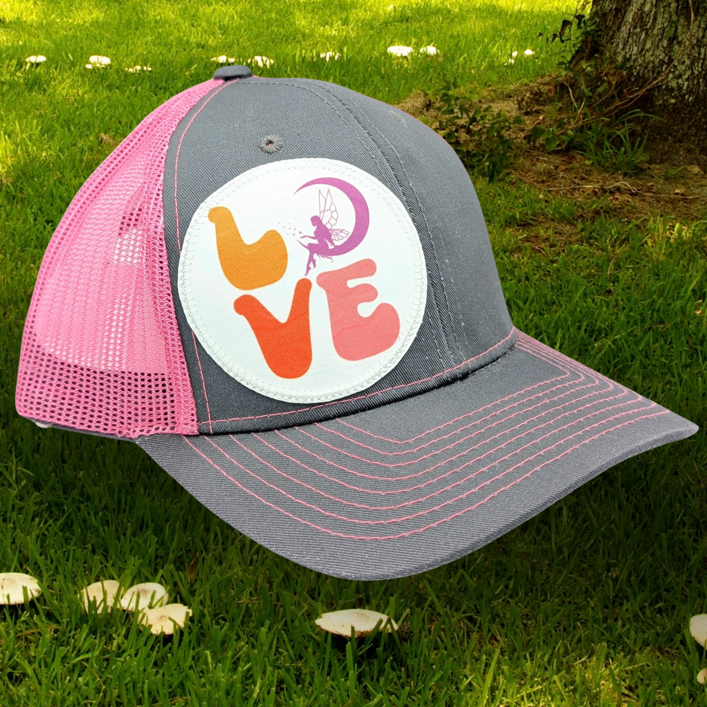 Front Right view Grey and Pink Trucker hat with hot pink stitching on the bill. Front and bill of hat are grey and the back is hot pink mesh. Large round white patch is on the right side of the hat. Patch has the word Love in bubble letters. The o is a crescent moon with a fairy sitting inside it.