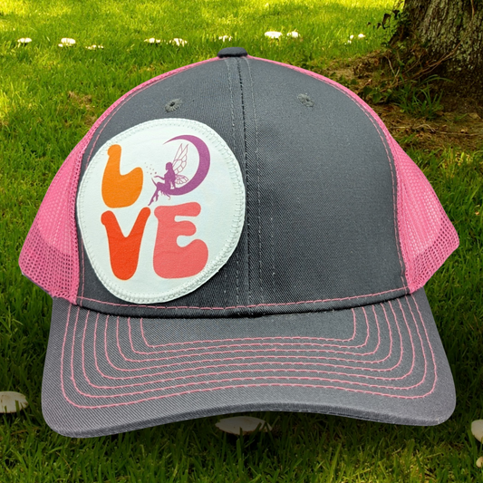 Front view of Grey and Pink Trucker hat with hot pink stitching on the bill. Front and bill of hat are grey and the back is hot pink mesh. Large round white patch is on the right side of the hat. Patch has the word Love in bubble letters. The o is a crescent moon with a fairy sitting inside it.