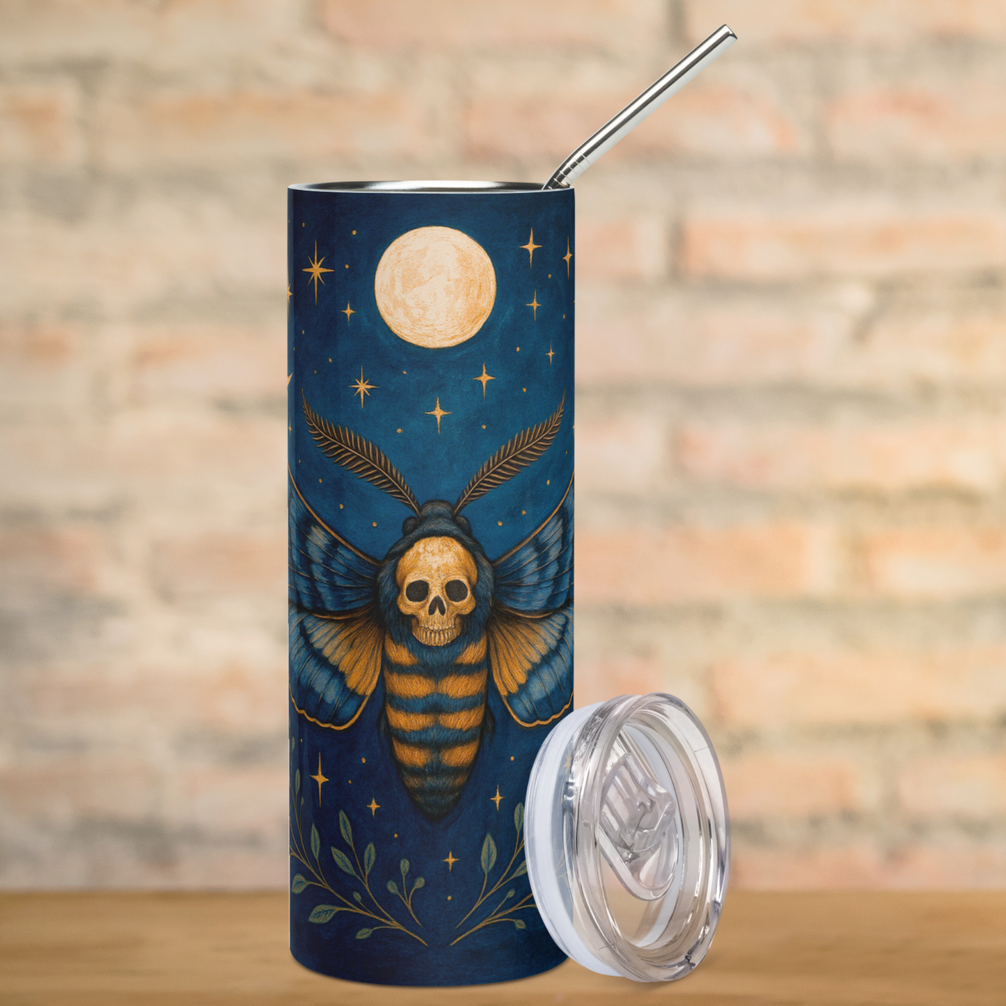 Death Head Moth Tumbler Stainless Steel 20 oz. View of front. Wings spread to each side with delicate feathered antennae the death head hawkmoth is shown in blues and golden browns with a gothic skull on the moth's back. Below the moth are spreading green branches. Above the moth is a full moon. Pagan Dark Cottagecore Coffee Tumbler
