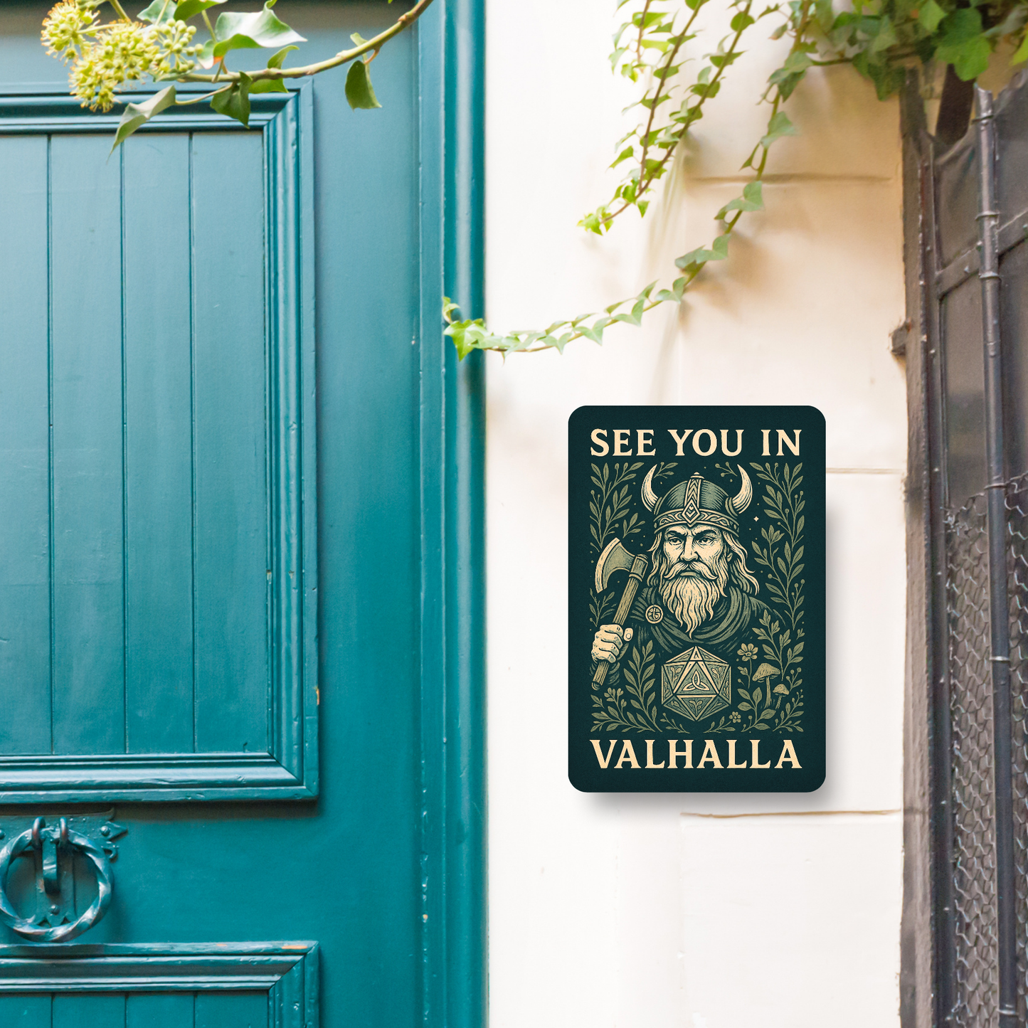 Metal Wall Poster 8x12 hanging next to a blue front door. Dark green Metal Sign with graphic in light golden browns. Bearded Viking wearing a horned helmet and holding an axe over his shoulder. D20 with symbols instead of numbers below the Viking. Botanical Design surrounding the image with mushrooms, vines, and flowers. At the top of the wall art it says, "See You In". At the bottom of the home decor, "Valhalla". Wall Art Apartment Decor