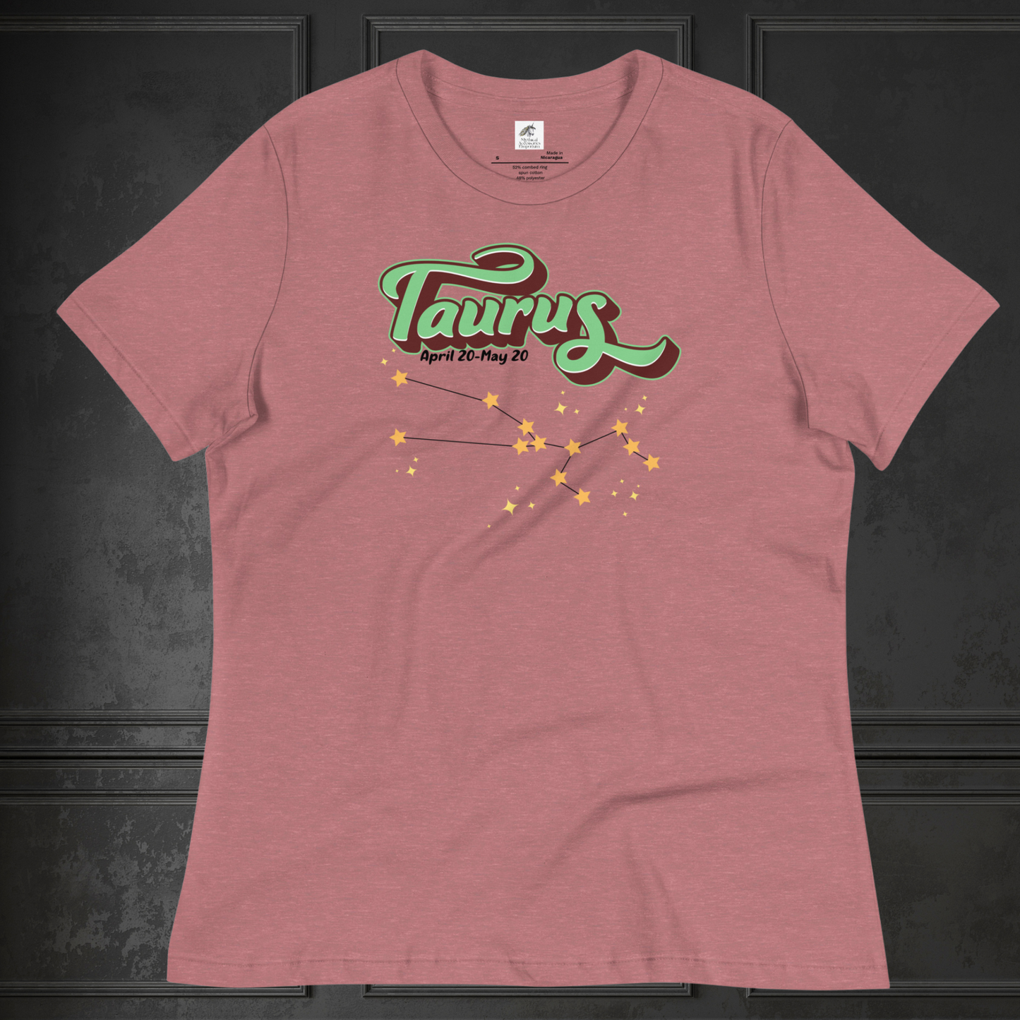 Front view of heather mauve Taurus Tee laying flat on a black surface. You can see the crew neck collar and short sleeves of the t shirt. This Astrology T-Shirt has the word Taurus in large leaf green script shadowed in deep brown for a 3-D effect. Below this it Says April 20-May 20. At the bottom is the star constellation for Taurus with yellow stars connected by a black line and smaller stars surrounding the constellation. The perfect April or May Birthday Gift for Her or yourself.