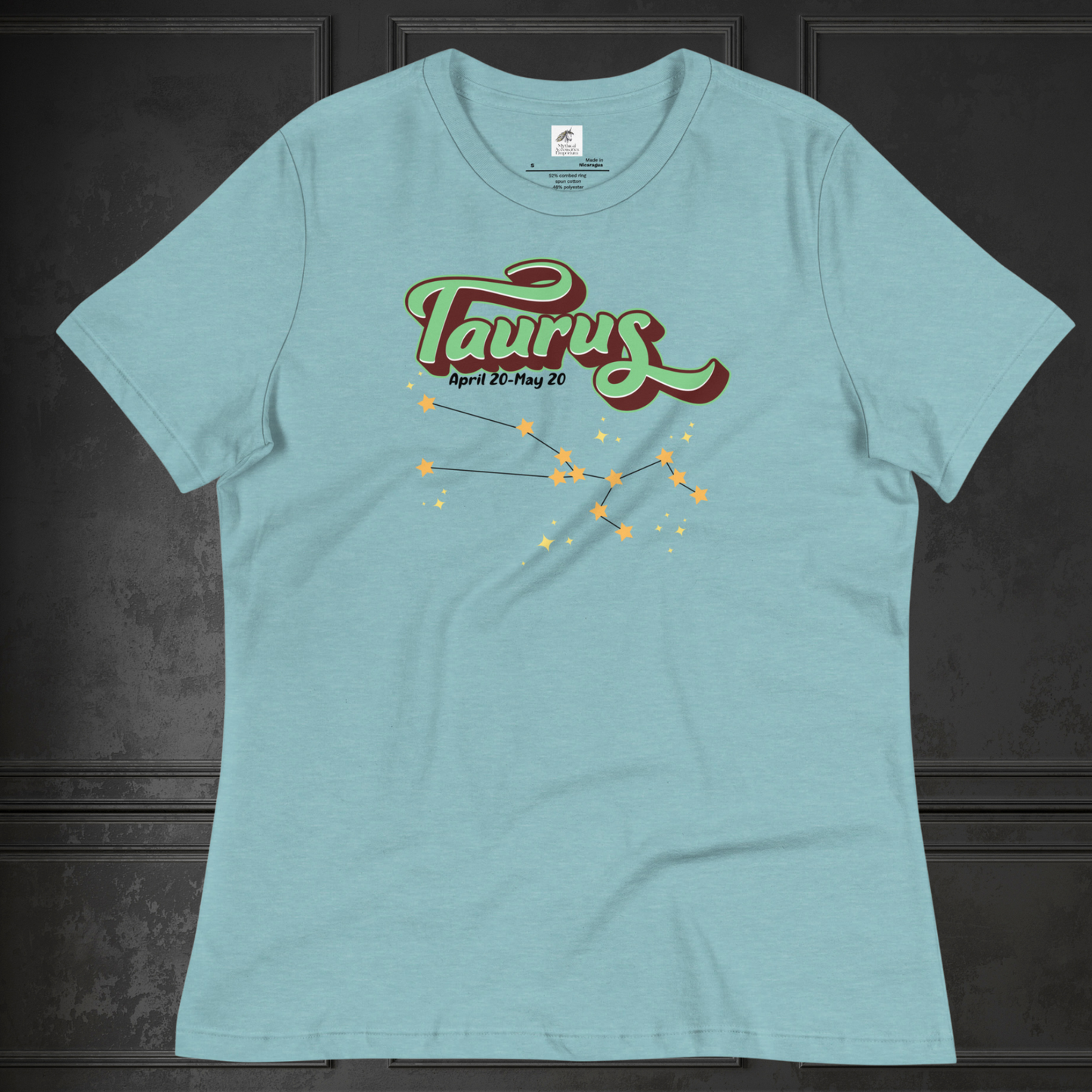 Front view of heather blue lagoon Taurus Tee laying flat on a black surface. You can see the crew neck collar and short sleeves of the t shirt. This Astrology T-Shirt has the word Taurus in large leaf green script shadowed in deep brown for a 3-D effect. Below this it Says April 20-May 20. At the bottom is the star constellation for Taurus with yellow stars connected by a black line and smaller stars surrounding the constellation. The perfect April or May Birthday Gift for Her or yourself.
