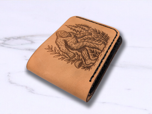Handmade BigFoot Leather Wallet. This dad style bi fold wallet is made out of veg tan leather. Laying on its back on a white surface this light brown wallet has a laser engraved Big Foot on the front walking through a pine forest and surrounded by vines with mushrooms on the path. The wallet is turned to see the folded edge of the leather.