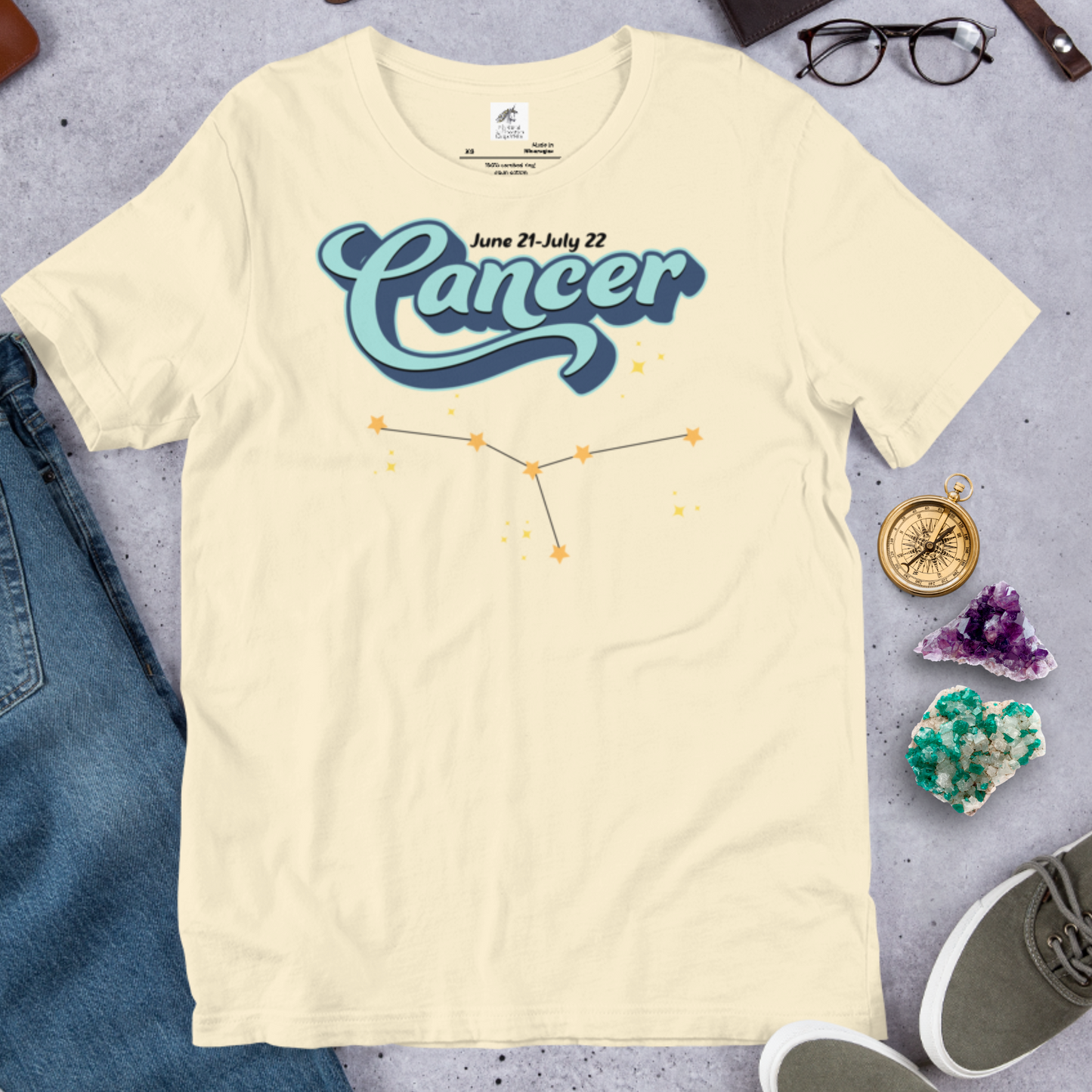 Front view of flat shirt natural color men's Cancer Tee. This astrology Cancer T-Shirt has the word Cancer in large light blue script shadowed in deep blue for a 3-D effect. Above this it Says June 21-July 22. Under Cancer is the zodiac star constellation with yellow stars connected by a black line and lighter yellow smaller stars surrounding this. The perfect June or July Birthday Gift for him or yourself.