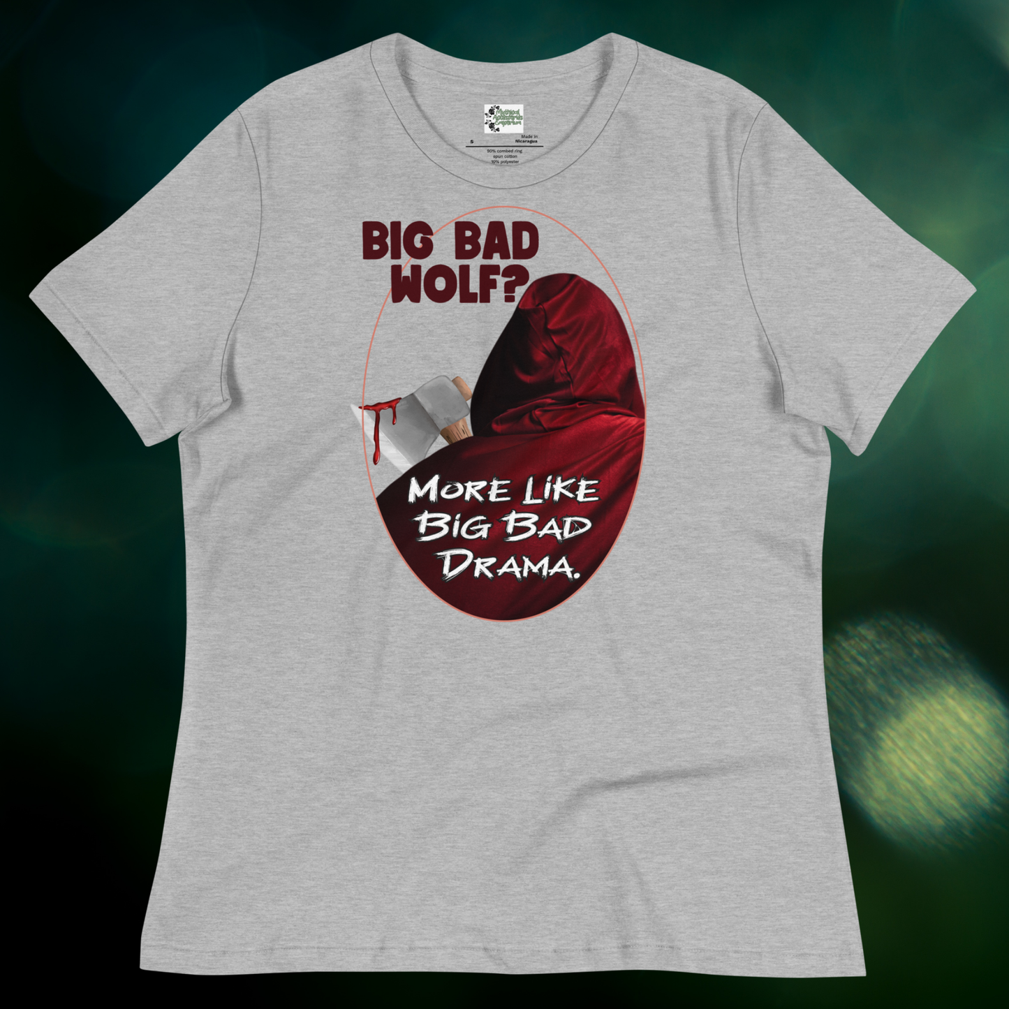Red Riding Hood Women's Dark Fantasy T-Shirt. Front View athletic grey Graphic Tee against a dark green background. Oval frame with thin red border. You can see the back of red riding hood with hood up. Bloody axe over her shoulder. Text Big Bad Wolf? More Like Big Bad Drama.