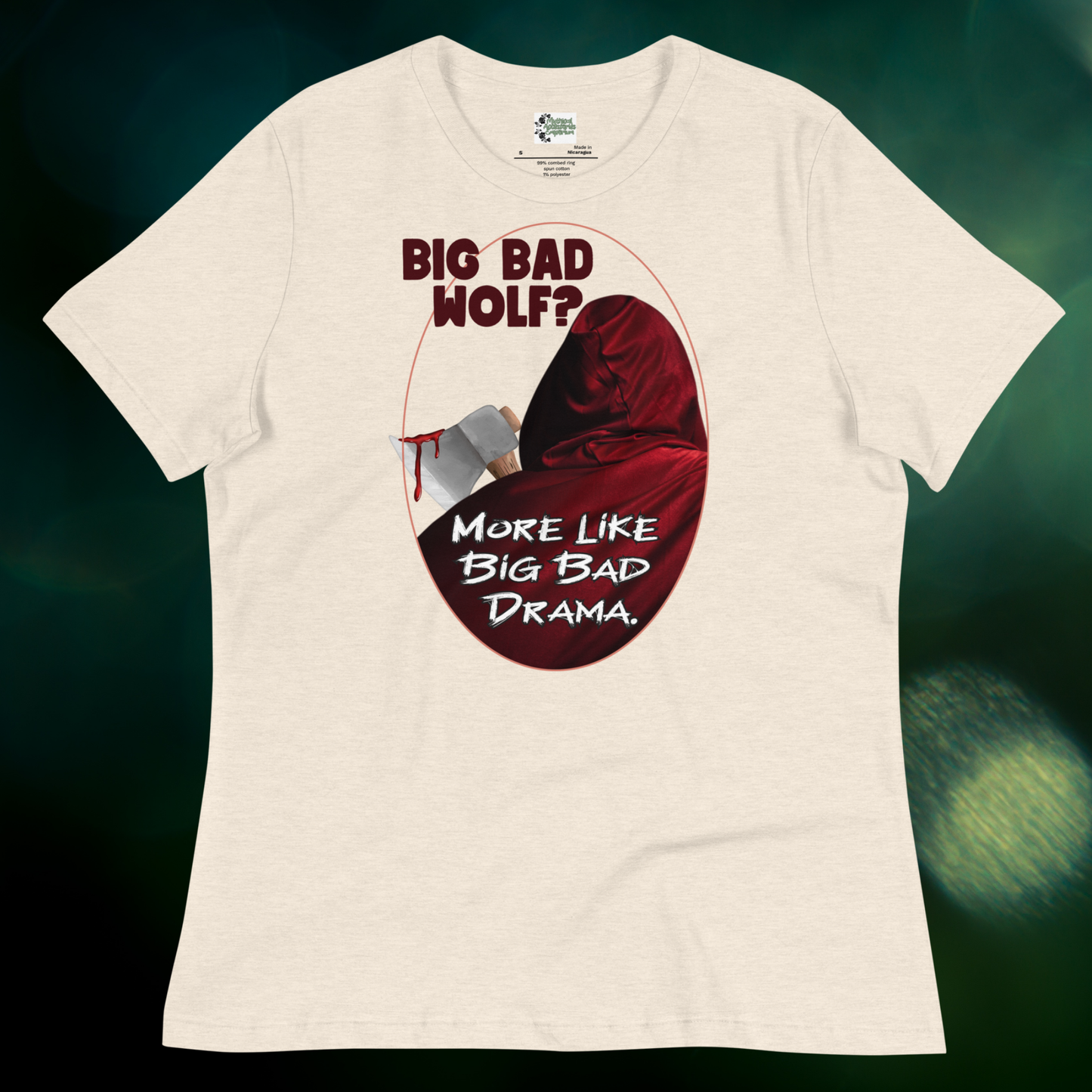 Red Riding Hood Women's Dark Fantasy T-Shirt. Front View heather prism natural Graphic Tee flat against a dark green background. This is a light tan or natural color cotton graphic tee. Oval frame with thin red border. You can see the back of red riding hood with hood up. Bloody axe over her shoulder. Text Big Bad Wolf? More Like Big Bad Drama. Women's Feminist Bookish Shirt