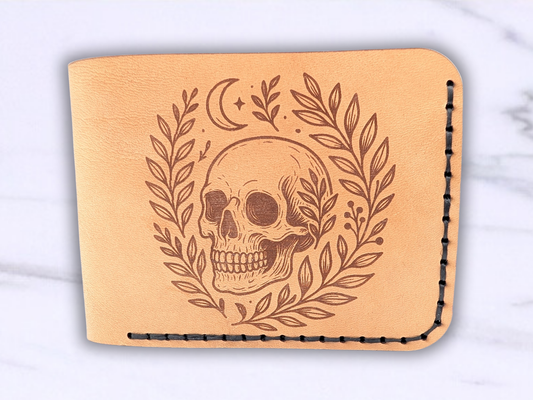 Front view of handmade leather bifold wallet with laser engraved skull. Front view of wallet with dark stitching on the right side and bottom. Leather is a light brown made from veg tan leather. Detailed skull with a crescent moon above it and vines surrounding both sides of the skull.