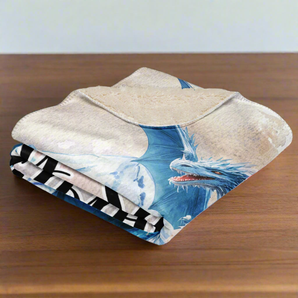 My Spirit Animal Dragon Blanket. Blanket is folded into a square, right side out, corner folded down to see natural sherpa. Blue Ice Dragon with wings spread standing in front of snow capped mountain range with a dragon silhouette flying above. Ice Dragon Fantasy Bookish Gift, Dungeons and Dragons DND Dungeon Master Gift, Fantasy Home Decor