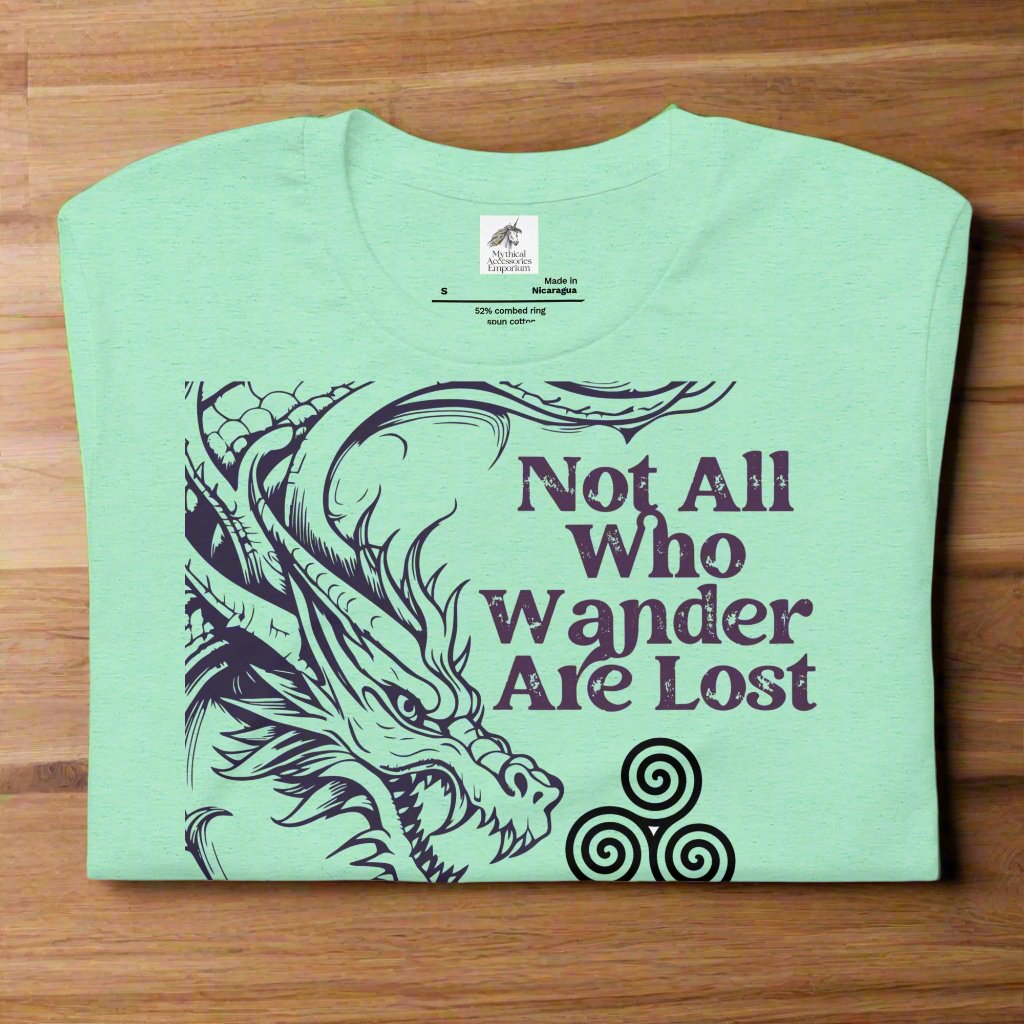 Heather mint men’s T-shirt, front view folded on counter. Right side features a dragon in smoky gray-purple with the Celtic triskele symbol emerging from its nostrils. Text above reads "Not All Who Wander Are Lost," with "Some Are Looking for Dragons" below.