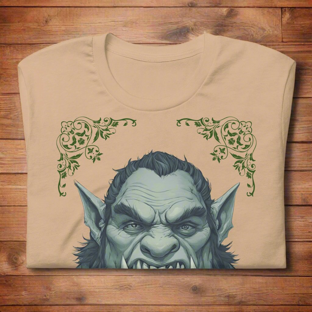 Front view of flat folded Men's Tan Graphic Tee with a black and white orc on the front. The orcs lower canines jut up as tusks over his top lip. He has a bulbous nose, eyebrows drawn down into a fierce expression, a large forehead, large pointed ears, his hair is black and pulled back with a widows peak, he has hair down the sides of his face and a medium sized beard. Framing the orc in four corners is 1920s retro floral border in dark green. This is a regular fit men's tshirt or a unisex shirt.