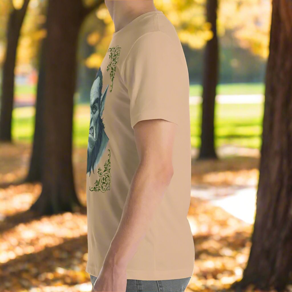 Left side view of Men's Tan Graphic Tee with a black and white orc on the front. The orcs lower canines jut up as tusks over his top lip. He has a bulbous nose, eyebrows drawn down into a fierce expression, a large forehead, large pointed ears, his hair is black and pulled back with a widows peak, he has hair down the sides of his face and a medium sized beard. Framing the orc in four corners is 1920s retro floral border in dark green. This is a regular fit men's tshirt or a unisex shirt.