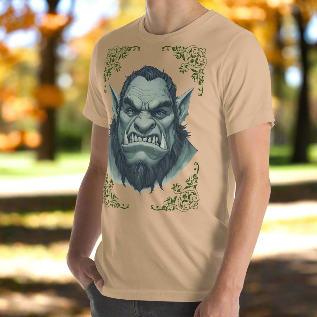 Front left side view of Men's Tan Graphic Tee with a black and white orc on the front. The orcs lower canines jut up as tusks over his top lip. He has a bulbous nose, eyebrows drawn down into a fierce expression, a large forehead, large pointed ears, his hair is black and pulled back with a widows peak, he has hair down the sides of his face and a medium sized beard. Framing the orc in four corners is 1920s retro floral border in dark green. This is a regular fit men's tshirt or a unisex shirt.