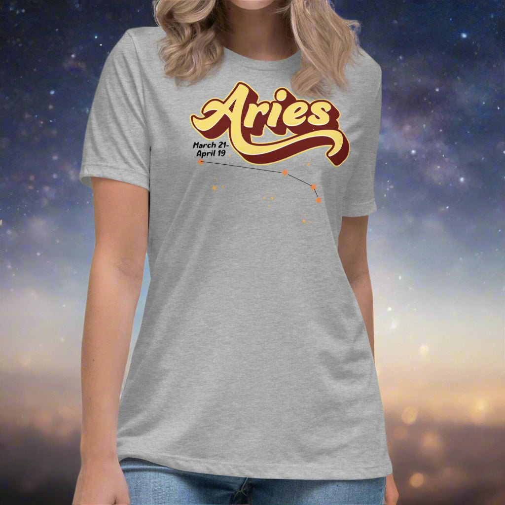 Woman wearing a athletic heather grey t-shirt facing forward. On the top is the word Aries in light yellow lined with maroon. Under the A are the dates March 21-April 19. Below his is the constellation Aries in bright yellow orange stars connected with a black line. Aries constellation zodiac astrology sign.
