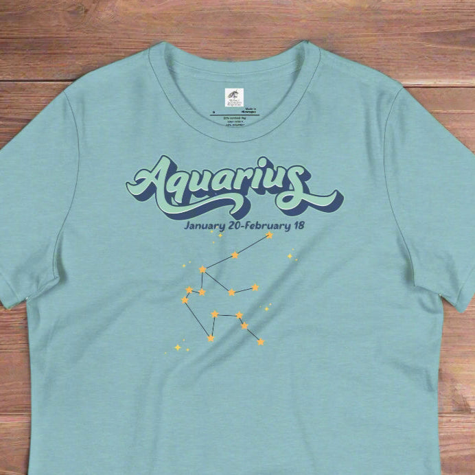 Flat lay heather blue lagoon astrology Aquarius women's T-shirt on a wooden counter. At the top in light blue is the word Aquarius outlined in deeper blue. Below this it says January 20- February 18. Under this is the Aquarius constellation with orange yellow stars connected by a black line. Astrology Zodiac Aquarius Woman's Tee