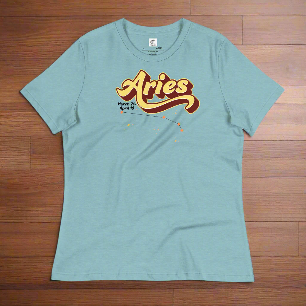 Aries Tee laying flat on a wooden counter, heather blue women's t-shirt. On the top is the word Aries in light yellow lined with maroon. Under the A are the dates March 21-April 19. Below his is the constellation Aries in bright yellow orange stars connected with a black line. Aries constellation zodiac astrology sign.