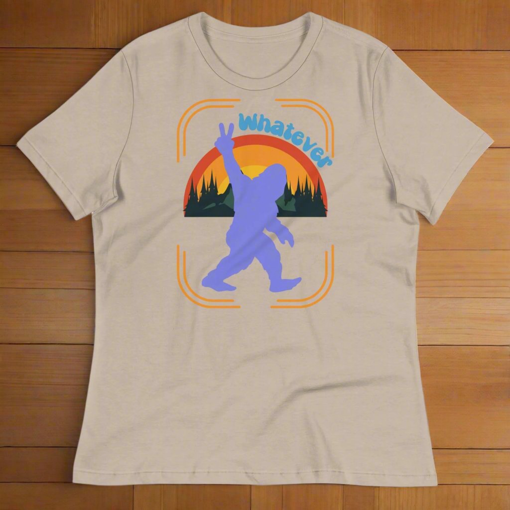 Heather Stone (beige-grey) Sasquatch Women's T-Shirt laying flat on a wooden surface. Big Foot in periwinkle against a rainbow and mountains showing a peace sign with text Whatever. Bigfoot Yeti Women's Tee