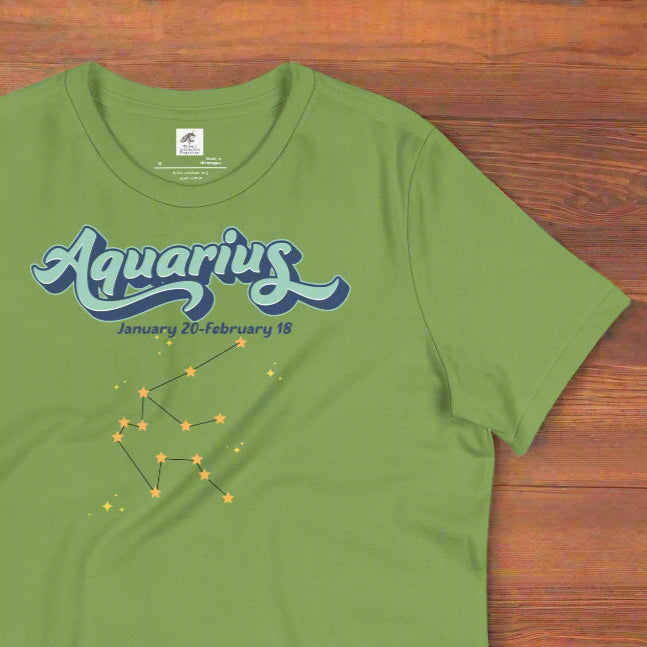 Flat lay leaf green astrology Aquarius women's T-shirt on a wooden counter. At the top in light blue is the word Aquarius outlined in deeper blue. Below this it says January 20- February 18. Under this is the Aquarius constellation with orange yellow stars connected by a black line. Astrology Zodiac Aquarius Woman's Tee