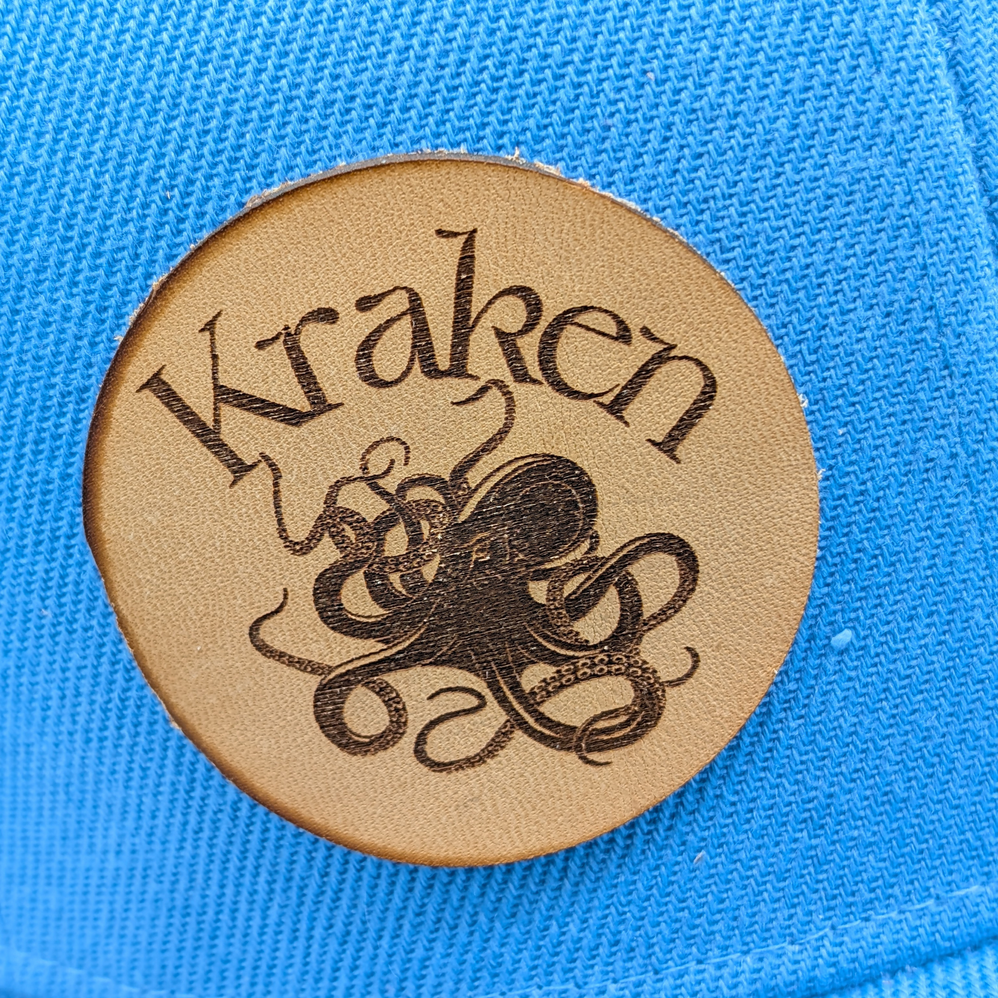 Up close view of Laser Engraved Leather Patch Text Kraken curved above the image of a Kraken. Kraken Viking Hat 