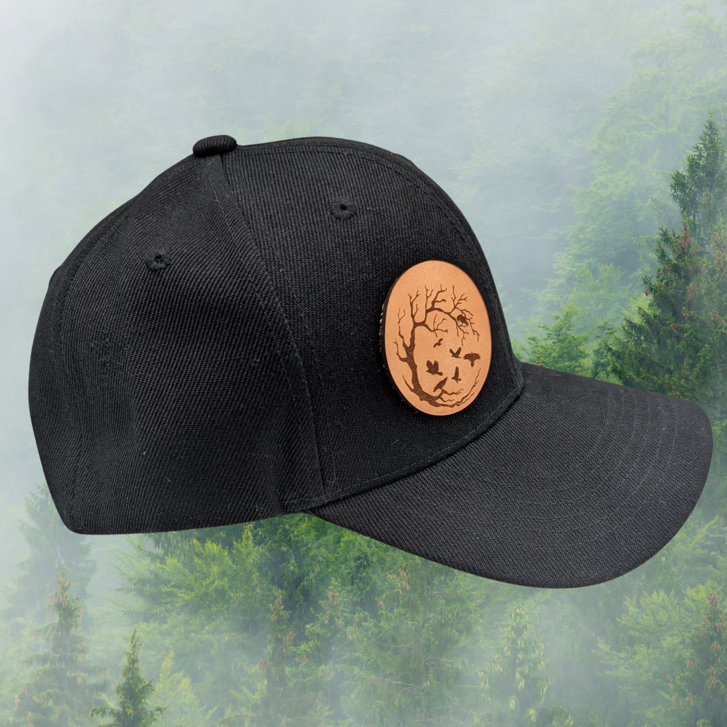 Right side view of Black Snapback Trucker Hat with a round leather laser engraved patch on the right side of the front. Leather Patch has a circle frame of a leafless tree in a with the branches reaching from the left to the right to make a circle. Ravens are in the branches and flying in the middle of the patch.