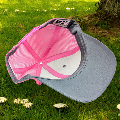 Bottom inside view of Grey and Pink Trucker hat with hot pink stitching on the bill. Front and bill of hat are grey and the back is hot pink mesh. Large round white patch is on the right side of the hat. Patch has the word Love in bubble letters. The o is a crescent moon with a fairy sitting inside it. 