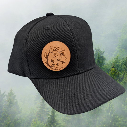 Front Right side view of Black Snapback Trucker Hat with a round leather laser engraved patch on the right side of the front. Leather Patch has a circle frame of a leafless tree in a with the branches reaching from the left to the right to make a circle. Ravens are in the branches and flying in the middle of the patch.