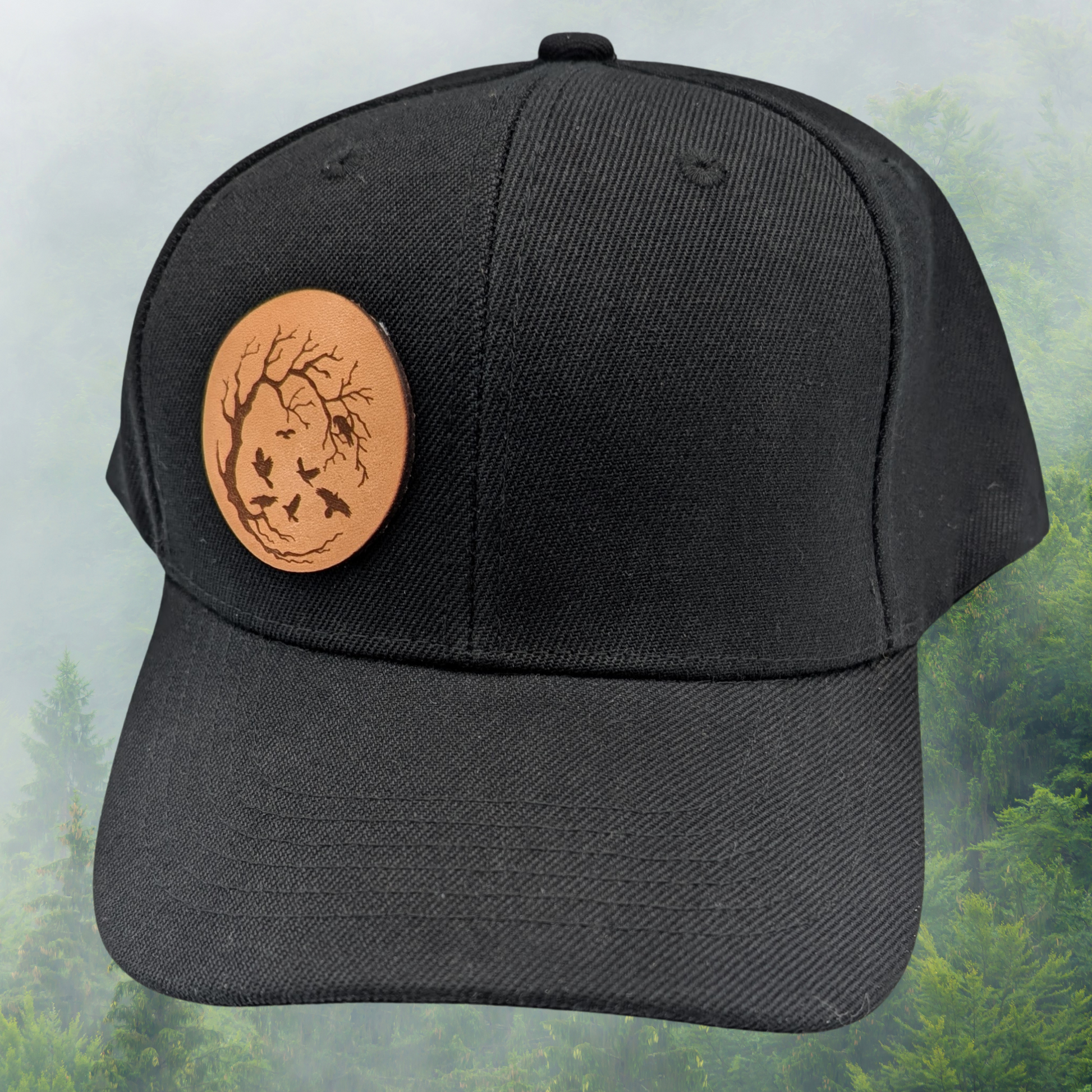 Front view of Black Snapback Trucker Hat with a round leather laser engraved patch on the right side of the front. Leather Patch has a circle frame of a leafless tree in a with the branches reaching from the left to the right to make a circle. Ravens are in the branches and flying in the middle of the patch.