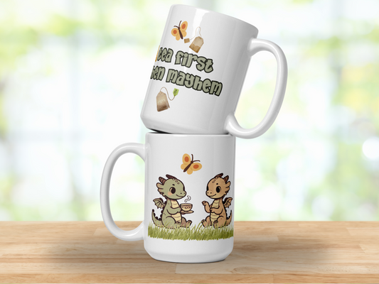 Ceramic 15 oz Mug. Two mugs stacked on top of each other so that you can see the front and the back. The bottom mug shows two kawaii chibi dragons facing each other drinking out of mugs. The top mug says "tea first then mayhem".