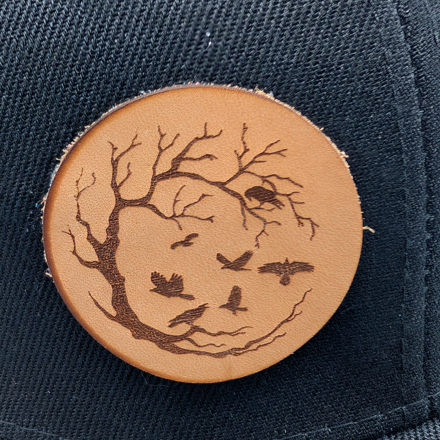 Up close view of the Leather Patch has a circle frame of a leafless tree in a with the branches reaching from the left to the right to make a circle. Ravens are in the branches and flying in the middle of the patch.