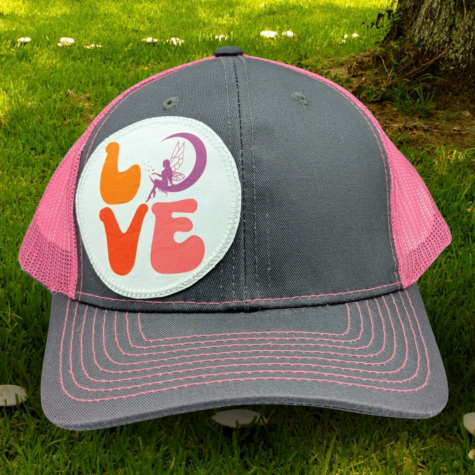 Front view of Grey and Pink Trucker hat with hot pink stitching on the bill. Front and bill of hat are grey and the back is hot pink mesh. Large round white patch is on the right side of the hat. Patch has the word Love in bubble letters. The o is a crescent moon with a fairy sitting inside it. 