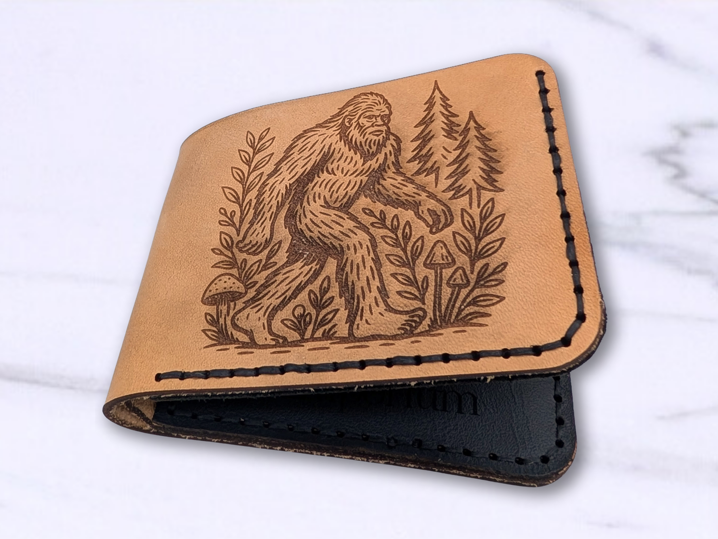 Handmade BigFoot Leather Wallet. This dad style bi fold wallet is made out of veg tan leather. Laying on its back on a white surface this light brown wallet has a laser engraved Big Foot on the front walking through a pine forest and surrounded by vines with mushrooms on the path. Stitching on the right and bottom in dark waxed cording. Wallet is slightly open to see the interior black pebbled card pocket with the same dark stitching. The double layers of the veg tan leather are visible stitched together