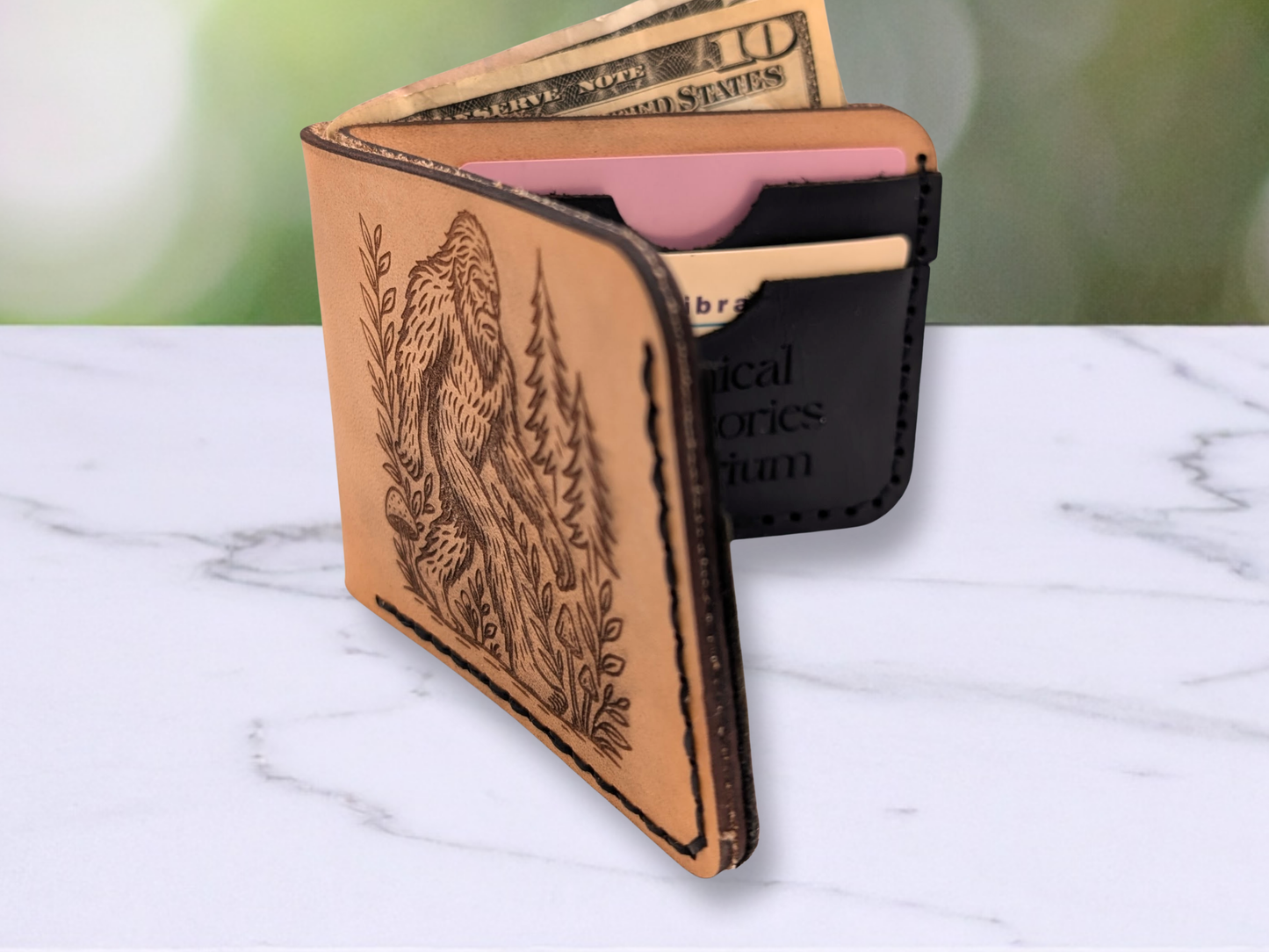 Handmade BigFoot Leather Wallet. This dad style bi fold wallet is made out of veg tan leather. Standin upback the light brown wallet has a laser engraved Big Foot on the front walking through a pine forest and surrounded by vines with mushrooms on the path. Stitching on the right and bottom in dark waxed cording. Wallet is slightly open to see the interior black pebbled card pockets with the same dark stitching. Card pockets have a credit card and a library card in them for sizing. 