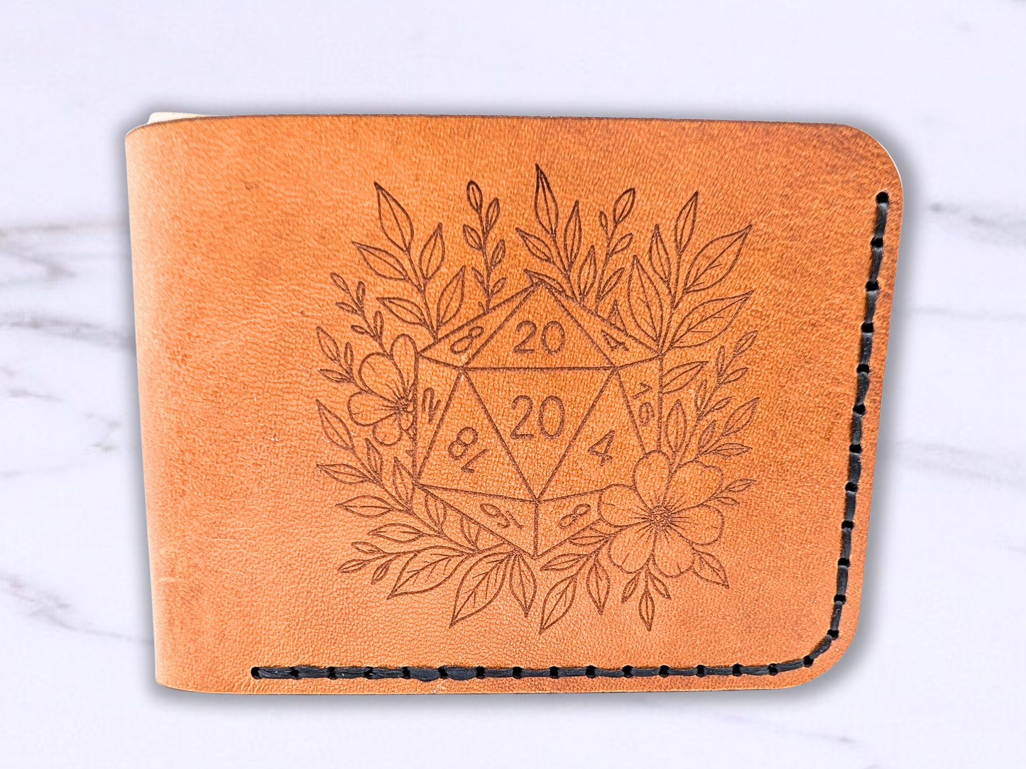 D20 veg tan wallet handmade with a D20 laser engraved on the front. The D20 is surrounded by leaves and a flower. This light brown leather bi fold wallet is dad style laying flat to get a front view. You can see the wallet has been stitched with black waxed cording. 