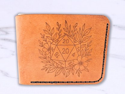 D20 veg tan wallet handmade with a D20 laser engraved on the front. The D20 is surrounded by leaves and a flower. This light brown leather bi fold wallet is dad style laying flat to get a front view. You can see the wallet has been stitched with black waxed cording. 