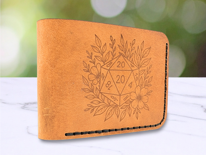 D20 veg tan wallet handmade with a D20 laser engraved on the front. The D20 is surrounded by leaves and a flower. This light brown leather bi fold wallet is dad style standing on the edge to showcase the folded side of the leather wallet with a view of the front engraving.