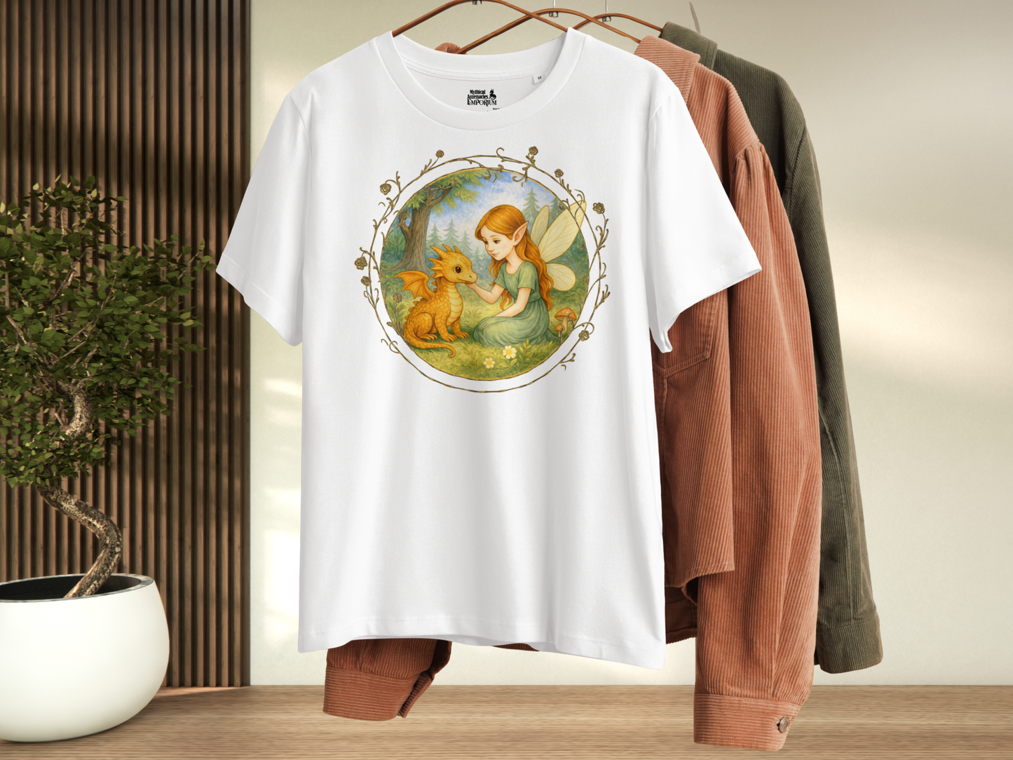 White t-shirt with a fantasy graphic hanging with other shirts. Circle shaped graphic is framed in gold rose vines. A fairy kneels in a forest meadow petting a reddish baby dragon. Fairy is wearing a long green dress and has auburn hair. Insect like wings. 