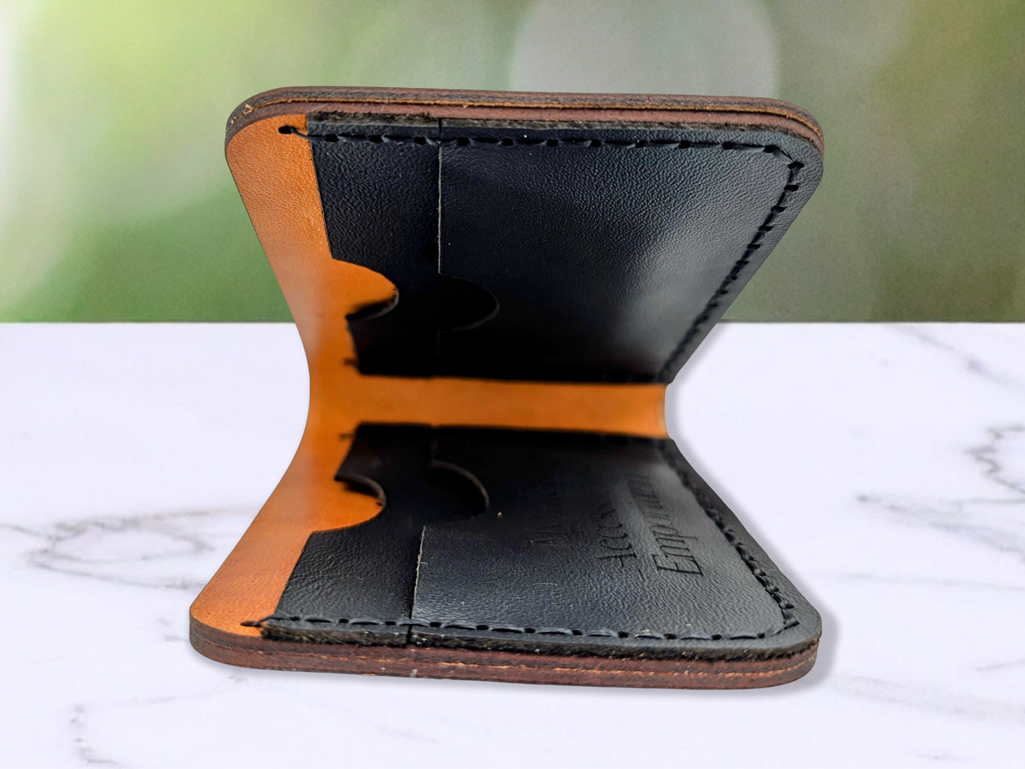 Edge View of the folded Leather Wallet to see into the wallet from the side. In this photo you see into the interior of the wallet to see all 4 card pockets made from black pebbled leather. The side on the counter has the logo engraved on the card pocket. There are two card pockets on each side of the wallet. Stitching is done in black waxed cord.
