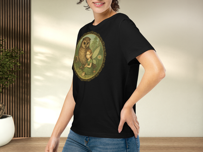 Person wearing a black t-shirt with a graphic design of a fairy and an owl. Left side view of model with hand on hip to show sleeve length and stretch.  Fairy is sitting on the ground in a forest next to a great horned owl. Fairy is wearing a long green dress with hair pulled back to show sharply pointed ears. 