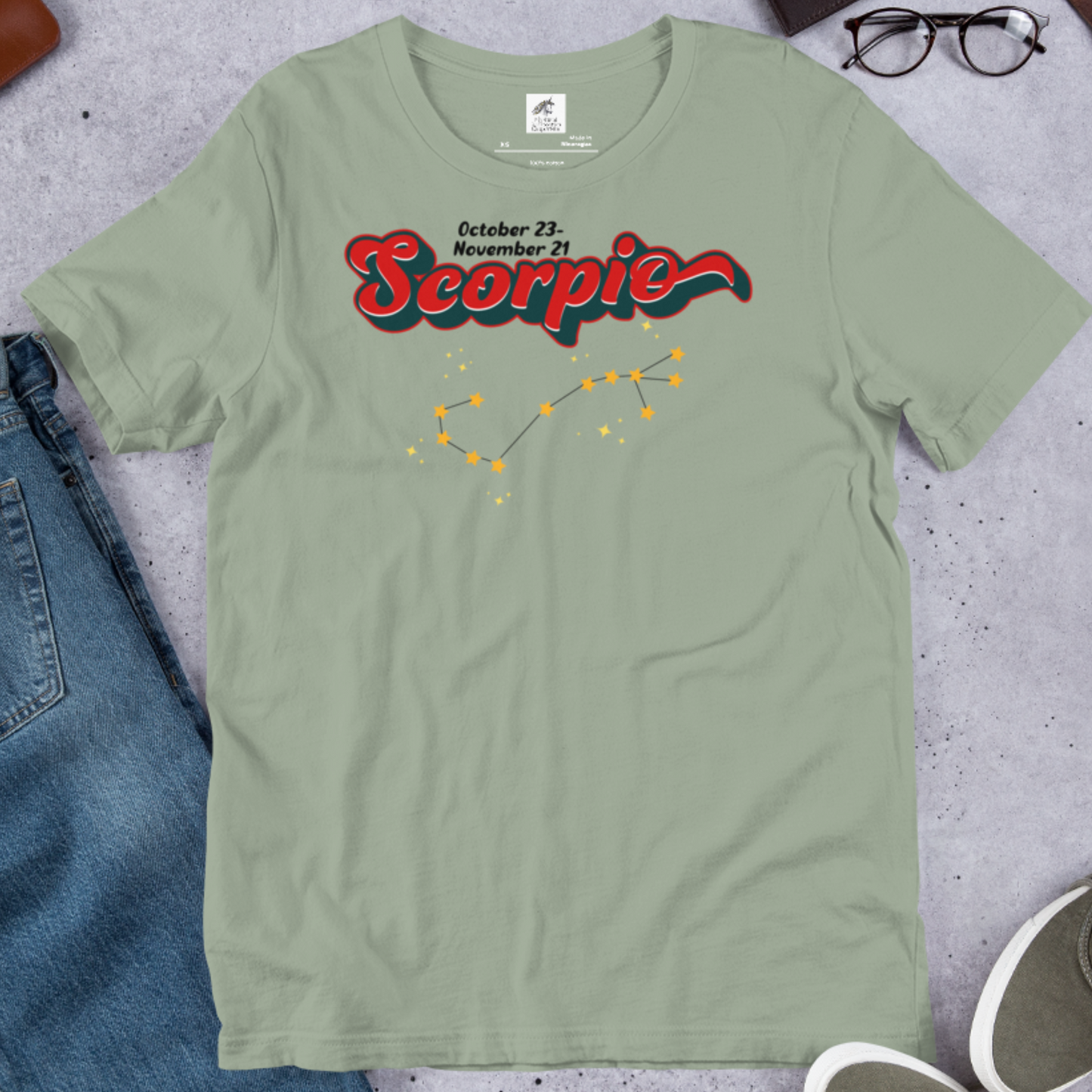Front view of flat sage men's Scorpio Tee. This astrology T-Shirt has the word Scorpio in large red script shadowed in black for a 3-D effect. Above  this it says October 23-November 21. Under Scorpio is the zodiac star constellation with yellow stars connected by a black line and lighter yellow smaller stars surrounding this. The perfect October or November Birthday Gift for him or yourself.