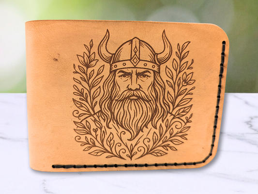 Front view Viking veg tan leather bi fold wallet. Laser engraved with a stern face of a Viking male warrior with a horned helmet, long beard and hair. He is surrounded with botanical vines on both sides. The laser engraving is a dark brown to black in color. The hand sewn stitching in dark waxed cord can be seen on the right and bottom of the wallet. The wallet has curved corners. 