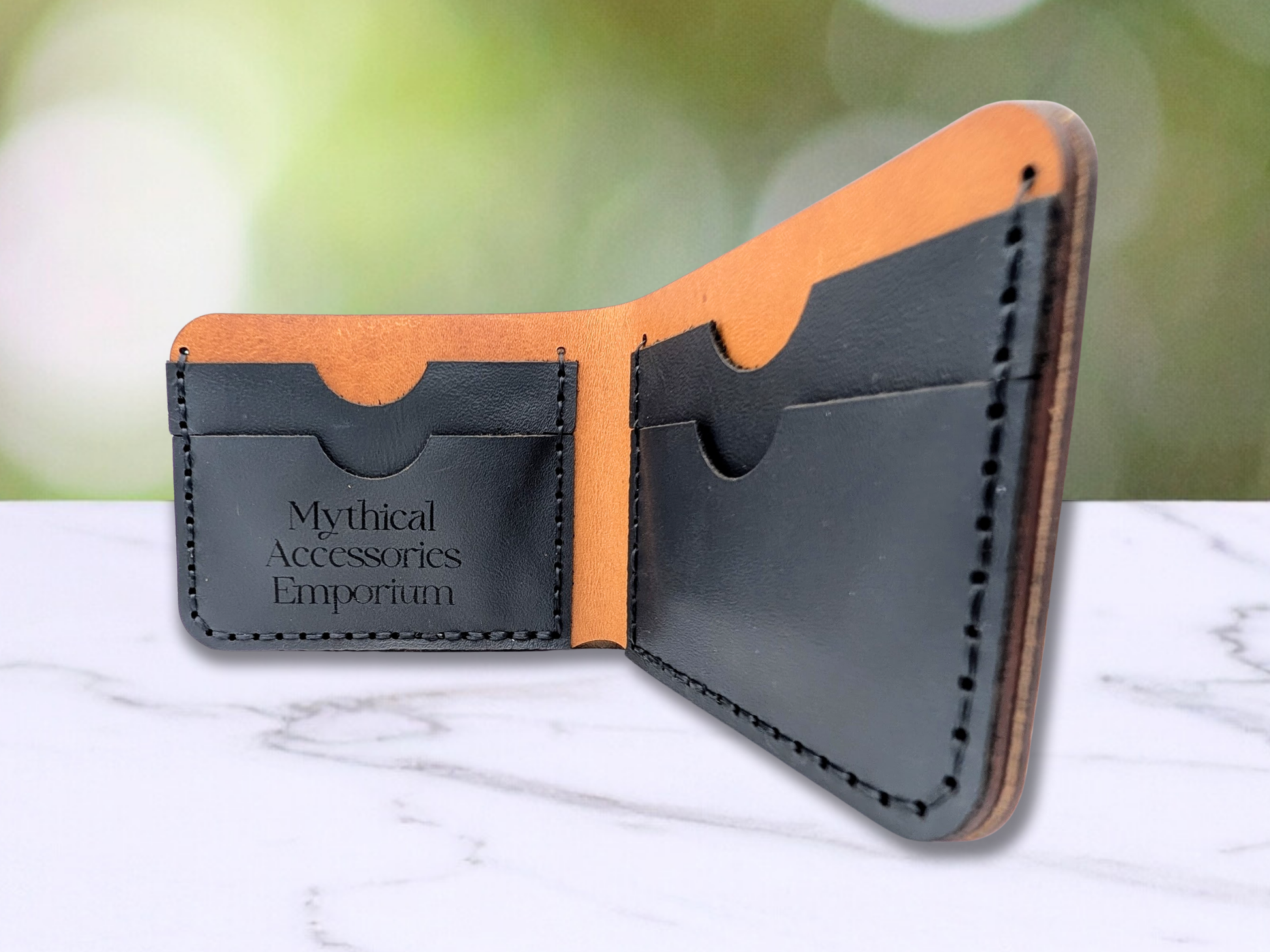 Handmade Leather Wallet open to the interior. There are two black pebbled leather card pockets on each side. They are sewn to the veg tan leather with black waxed cording. On one side of the wallet you can see the logo, Mythical Accessories Emporium has been laser engraved. Each card pocket has a rounded notch to allow cards to be removed easier.