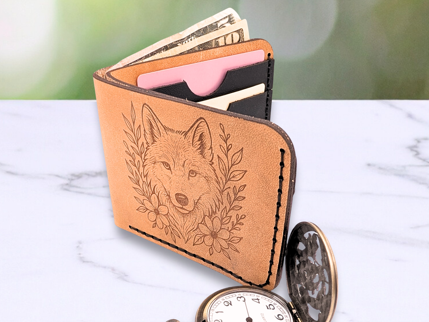 Leather veg tan wallet with laser engraved wolf head looking out at you on the front. To each side of the wolf are vines and flowers. In the photo you can see dark stitching, the cash compartment has bills that are pulled up to show off the opening for the cash between two pieces of light brown leather. The wallet is standing up opened and you glimpse the interior black pebbled leather card pockets with cards in them. The edges of the leather are dark along with the engraving. 