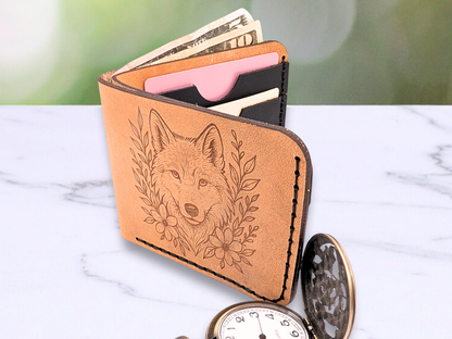 Leather veg tan wallet with laser engraved wolf head looking out at you on the front. To each side of the wolf are vines and flowers. In the photo you can see dark stitching, the cash compartment has bills that are pulled up to show off the opening for the cash between two pieces of light brown leather. The wallet is standing up opened and you glimpse the interior black pebbled leather card pockets with cards in them. The edges of the leather are dark along with the engraving. 
