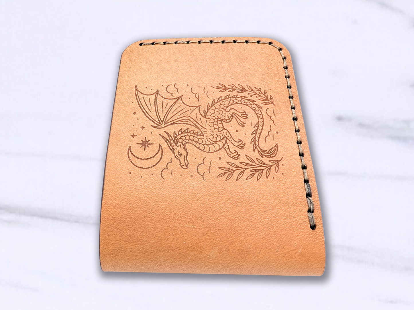 Leather dragon wallet laying flat on a white surface. The folded side of the wallet is facing you with the wallet sideways. Stitching and dragon image are visible. Photo shows the curve and color of the natural leather.