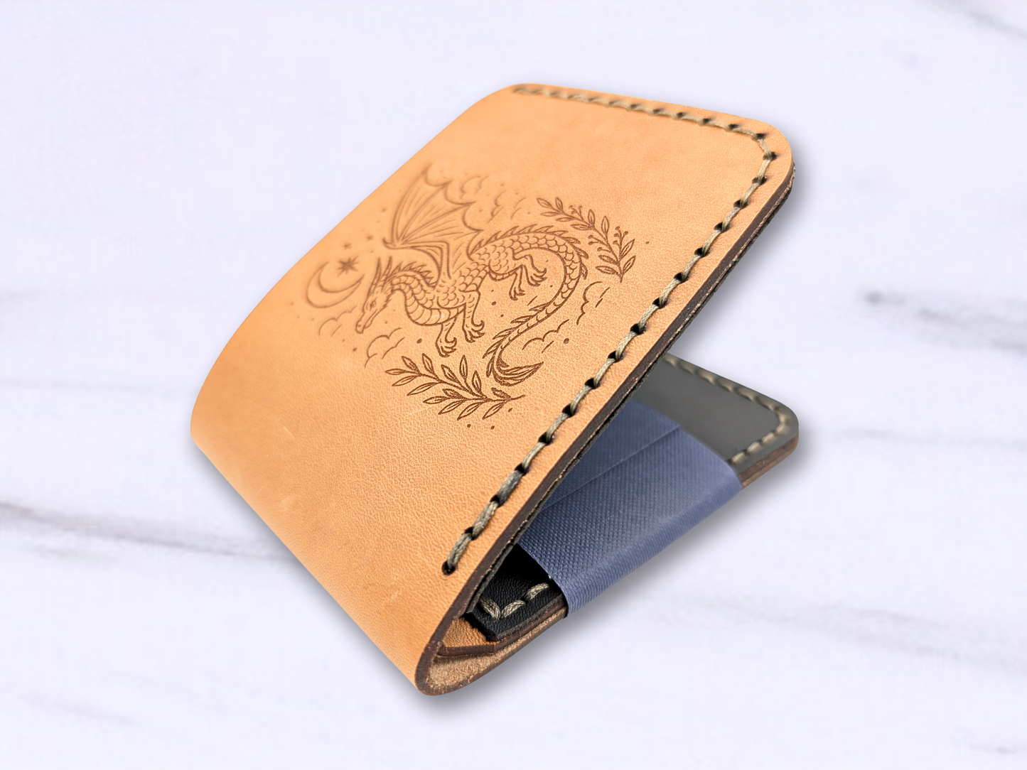 Wallet is laying on a white surface. the leather wallet is slightly open and you are looking at it from the bottom. You can see the stitching on both sides of the wallet, the dragon image on the front, and you glimpse the black leather card pockets on the interior. You see the darkened edges of the leather at the bottom of the cash compartment and a bit of the suede is visible inside the compartment at the fold. 
