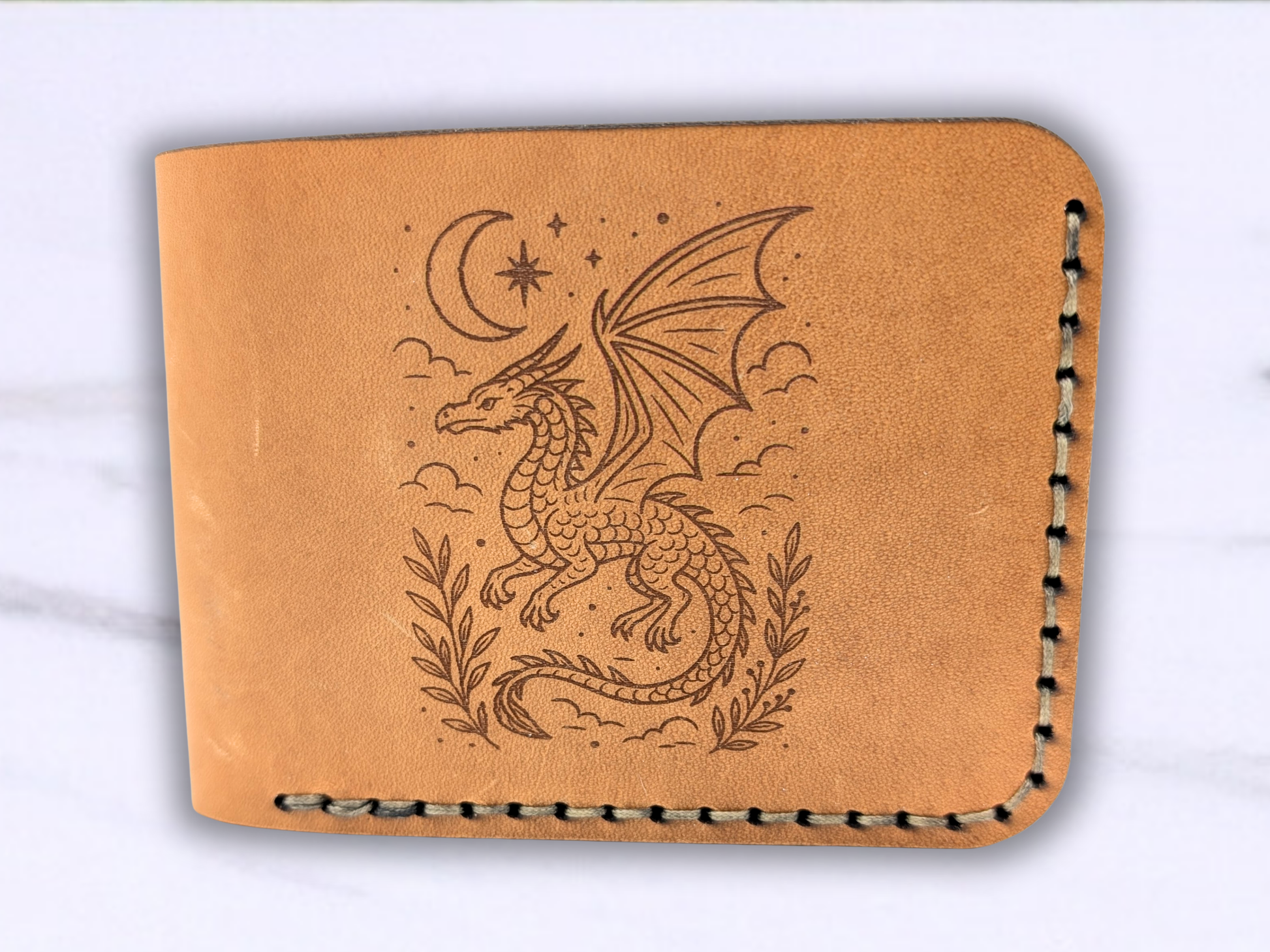 Front view of the folded bifold dragon leather wallet facing up. You see the stitching coming down the right side and the bottom of the wallet. The folded side is to the left. On the front is the laser engraved image of a flying dragon amongst the clouds with a crescent moon and a star above and below to each side is a vine of leaves. 